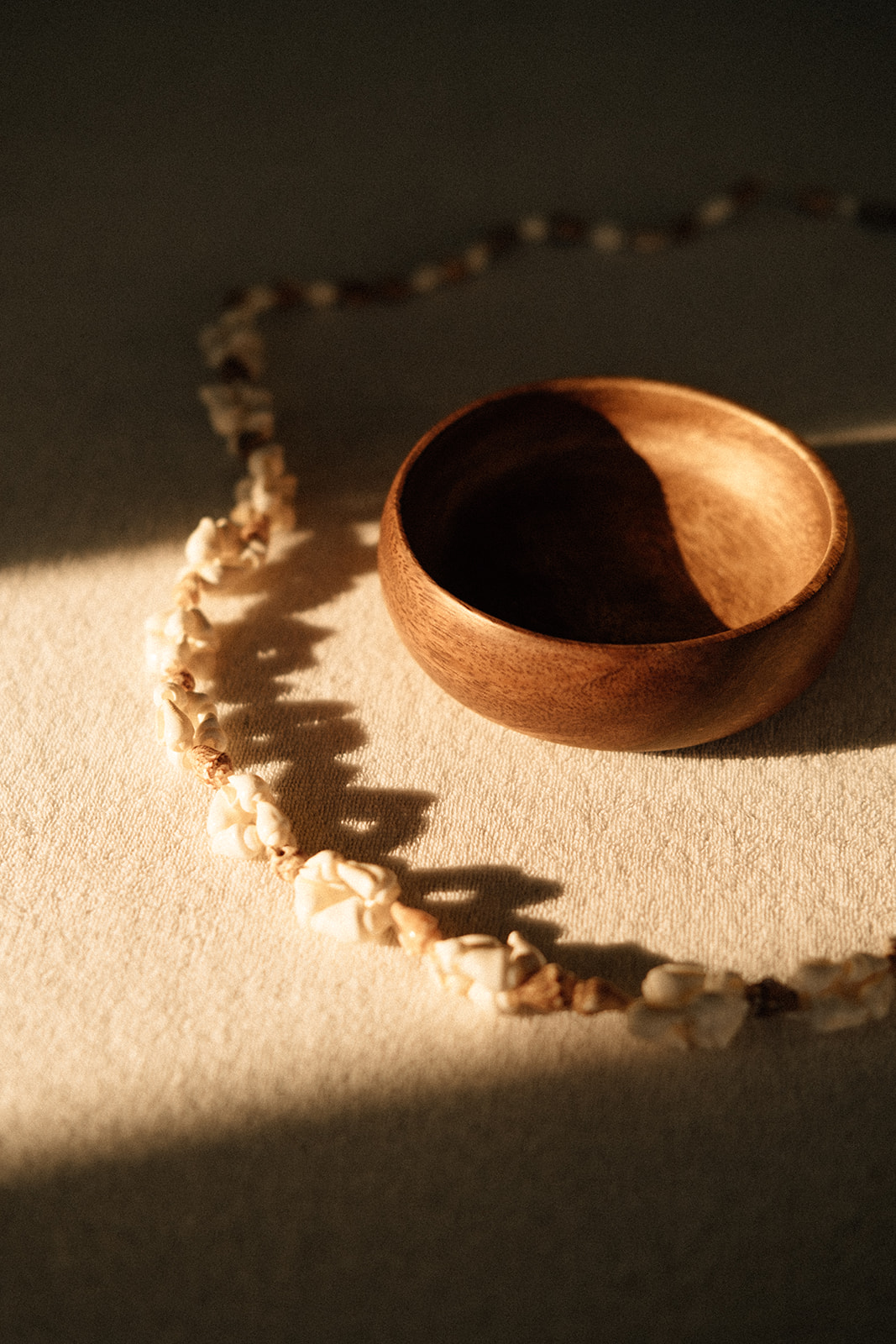 Eine leere Holzschale befindet sich auf einer Massageliege. Um die Schale befindet sich eine Muschelkette. Ein warmer Sonnenstrahl scheint von links oben auf die Schale und die Kette.