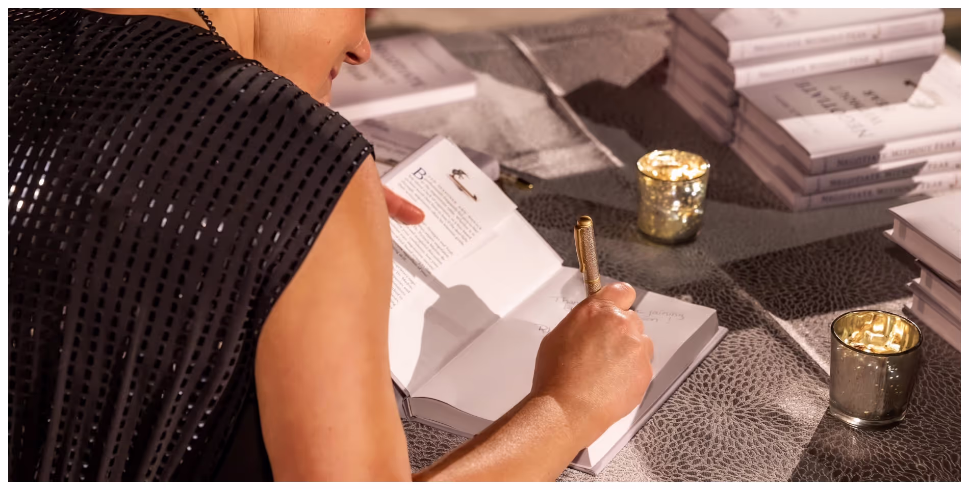 Person signing a book with a gold pen at a table with lit candles and stacks of books.