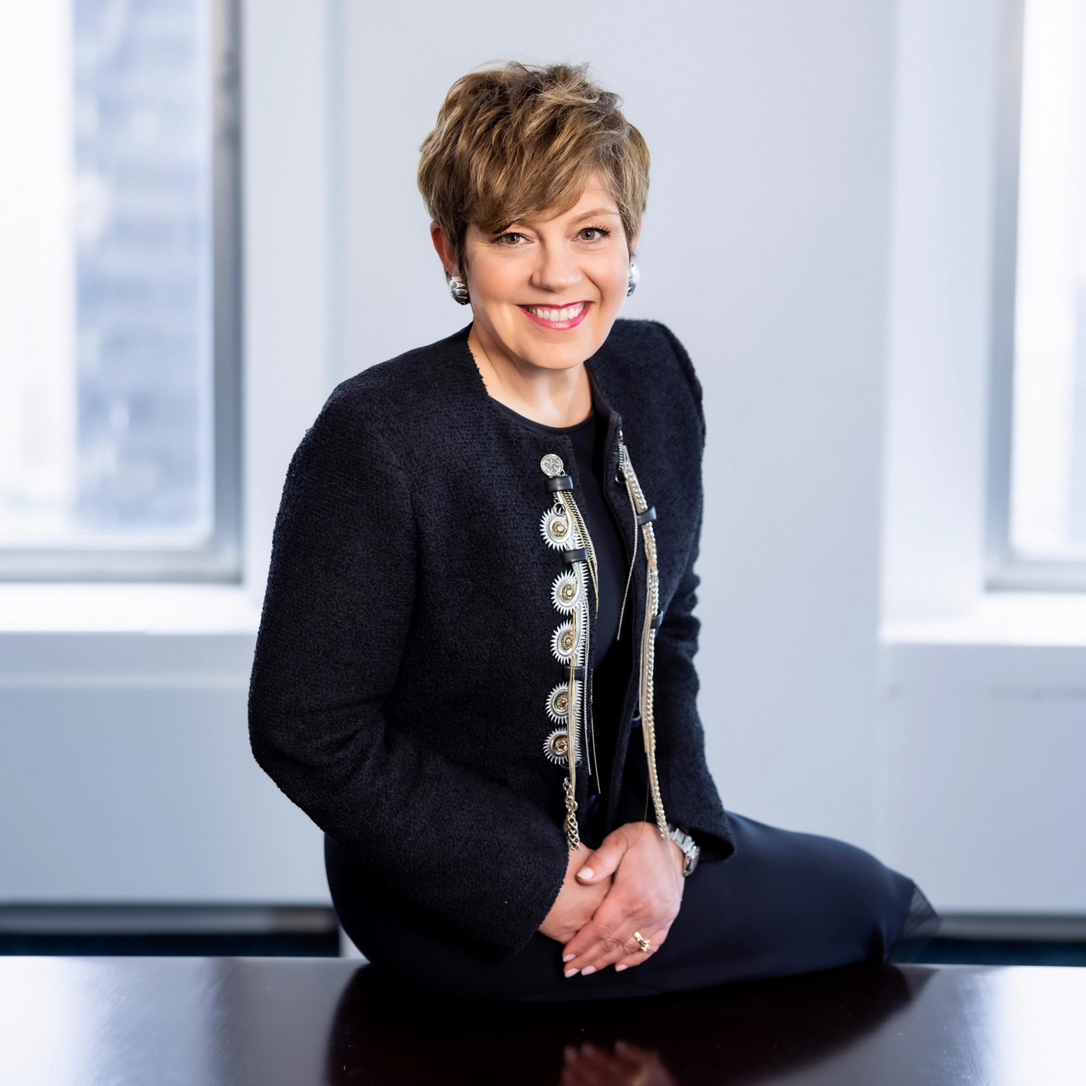Smiling woman with short hair wearing a black jacket sitting on a table indoors near windows.