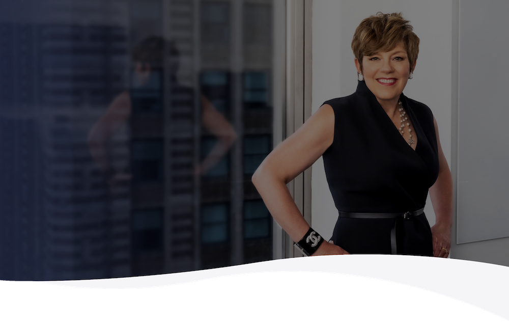 Smiling woman in black dress with pearl necklace standing by office window with cityscape view.