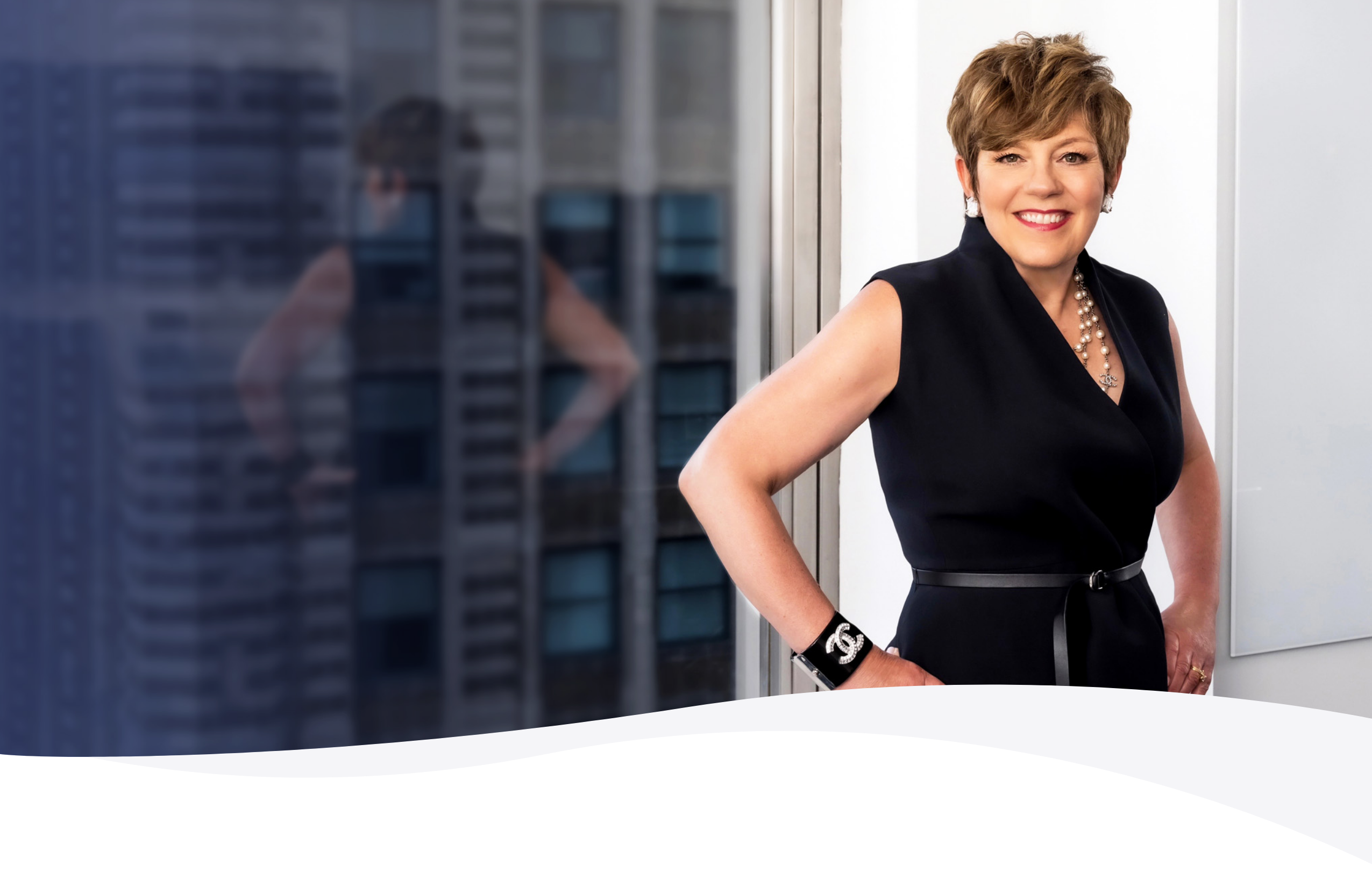 Victoria Medvec, a smiling woman with short hair in a sleeveless black dress, stands by a window with city view reflection.
