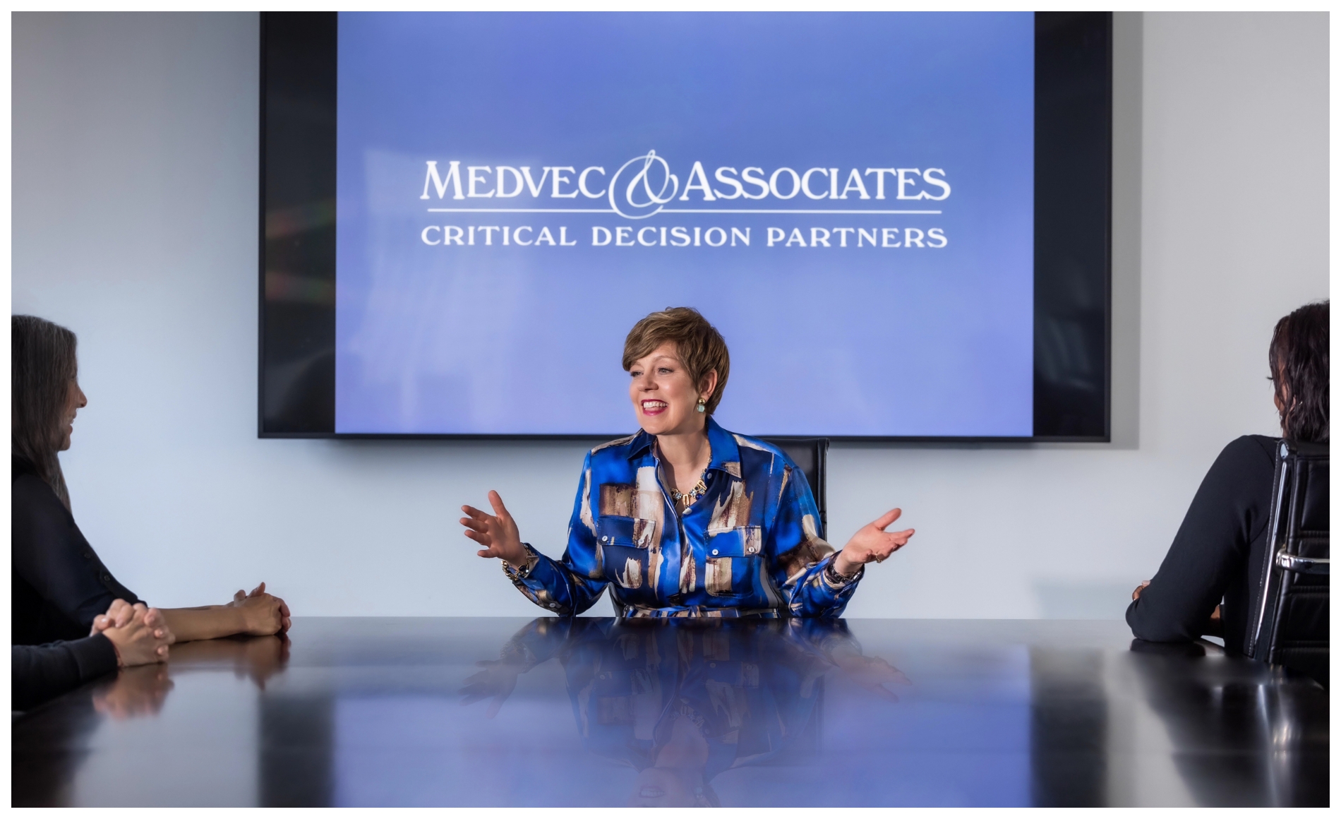Dr. Victoria Medvec, a middle-aged woman with short brown hair, communicates with her hands at the head of a conference table while others office workers look on.