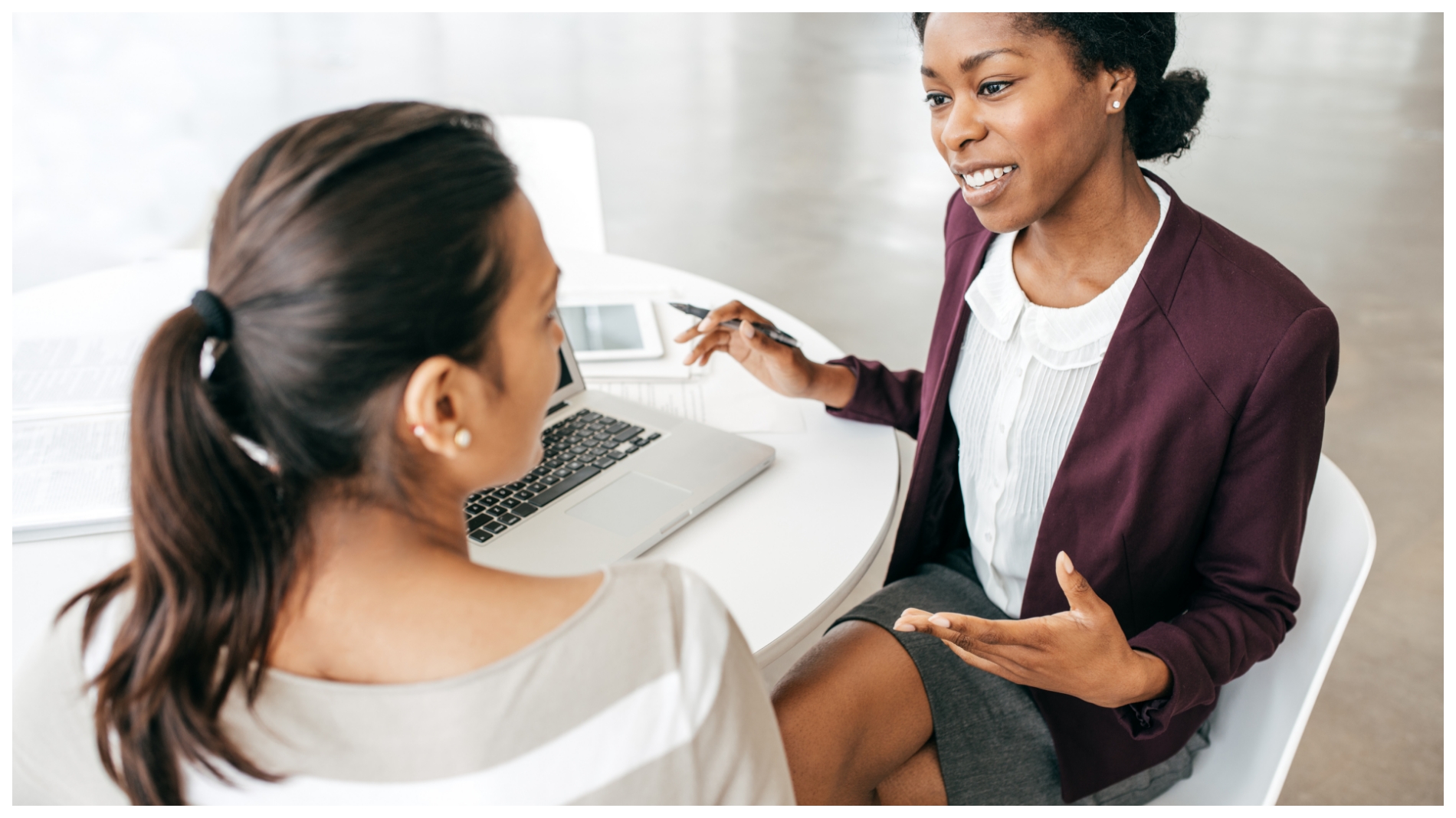Two women conversing at a table in an office with a laptop.