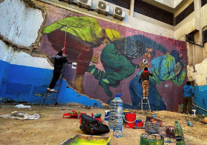Three artists painting a vast mural, there is lots of paint and water bottles on the ground.