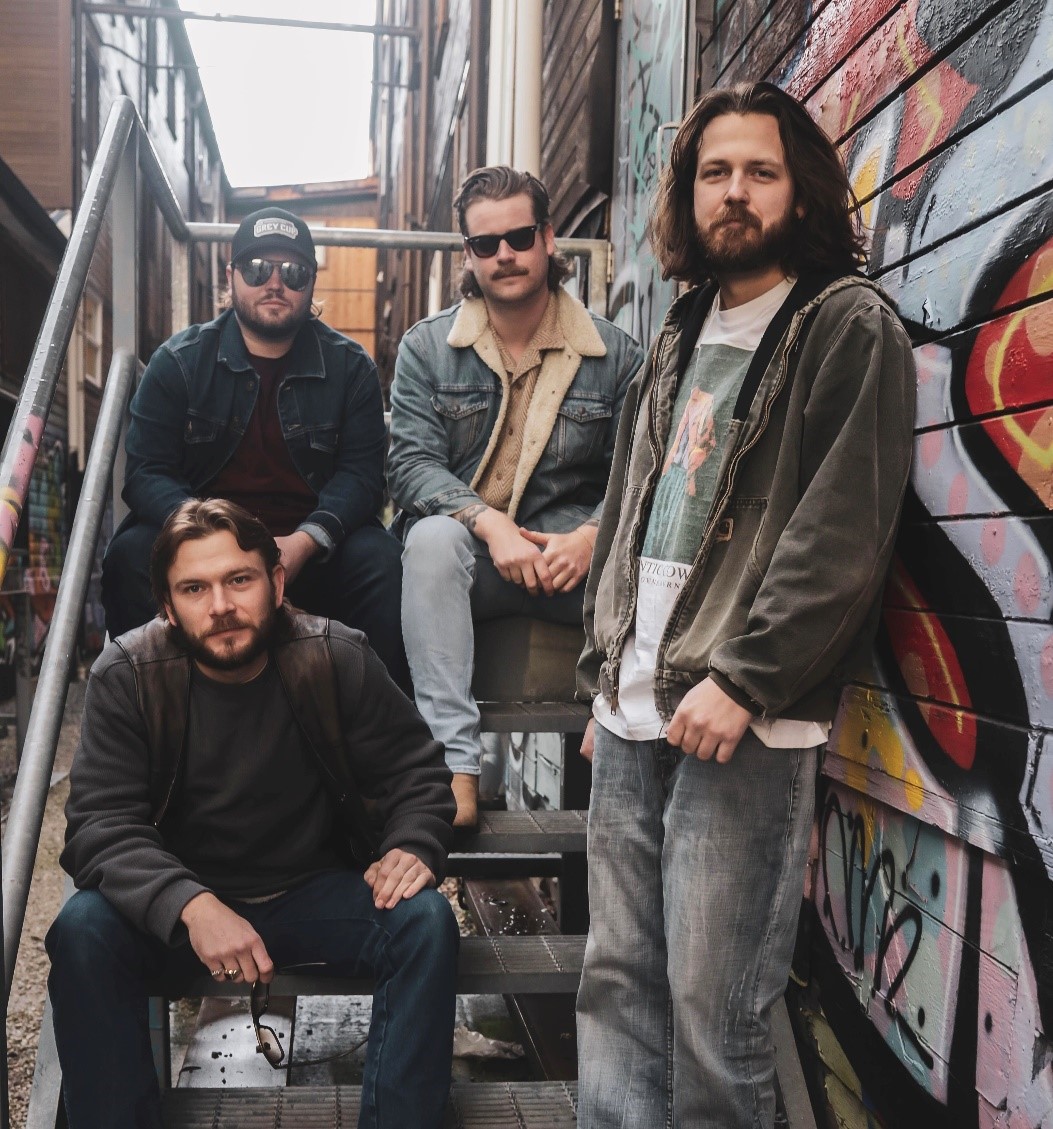 Rock band posing near a building with murals. One band member is standing while three other band members are sitting on the steps.