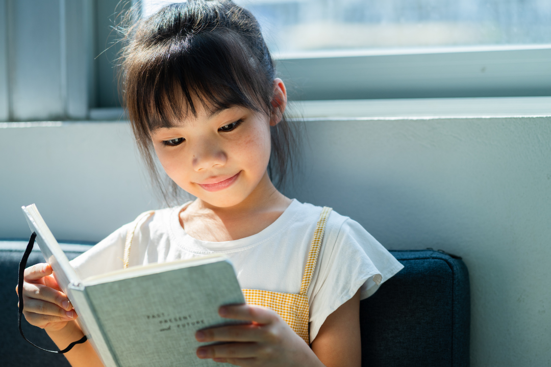 Smiling girl reading a book while sitting by a sunny window.