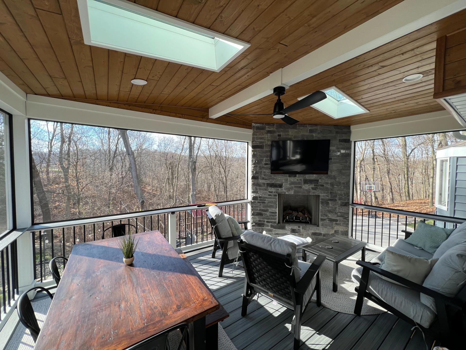 Covered outdoor deck with wooden ceiling, skylights, stone fireplace, mounted TV, cushioned seating, and a wooden dining table with chairs overlooking a wooded backyard.