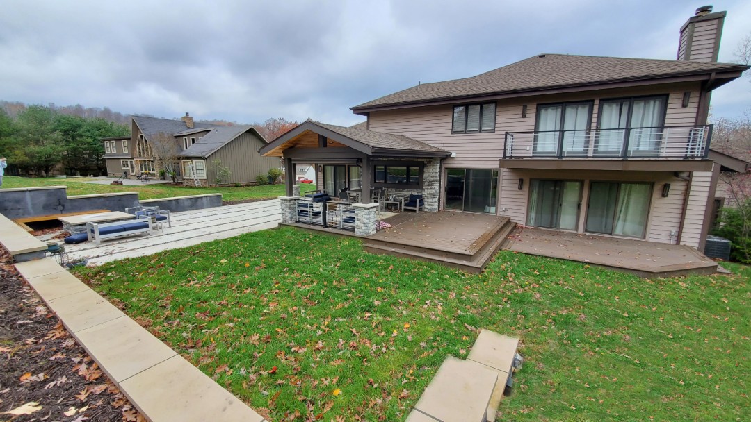 Two-story modern house with wooden siding, large deck, and covered patio surrounded by green lawn and autumn leaves under a cloudy sky.