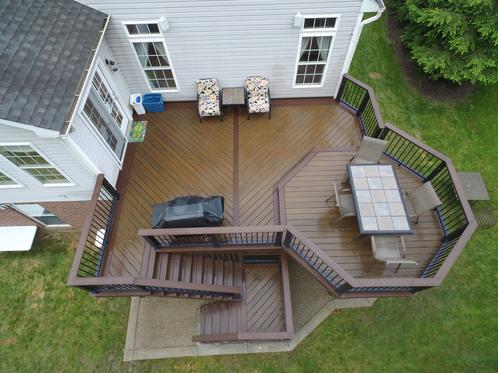 Aerial view of a composite backyard deck with stairs, outdoor seating, a covered grill, and green lawn surrounding it.
