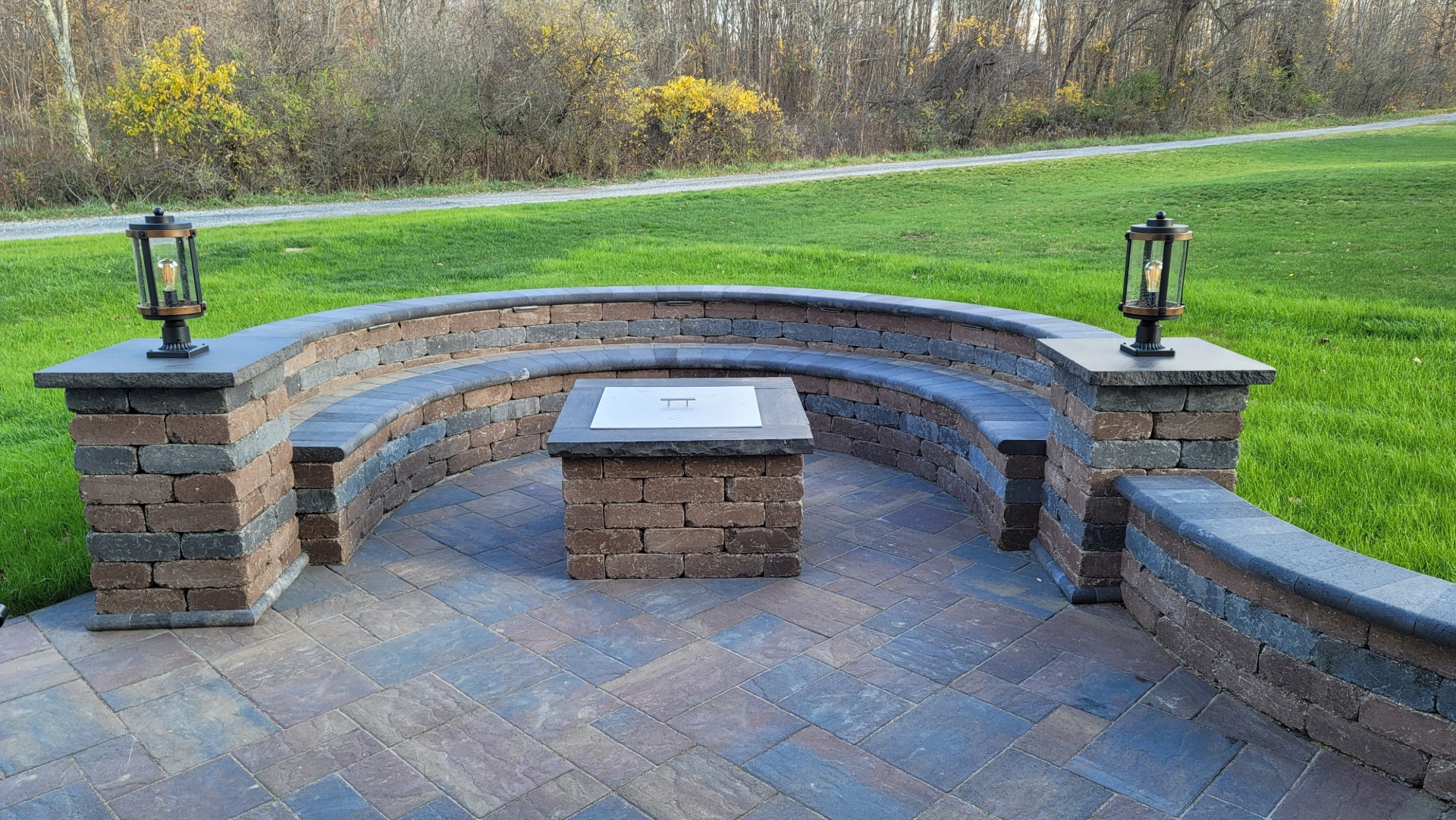 Curved stone patio bench with two lanterns and a square stone fire pit in a grassy outdoor setting.