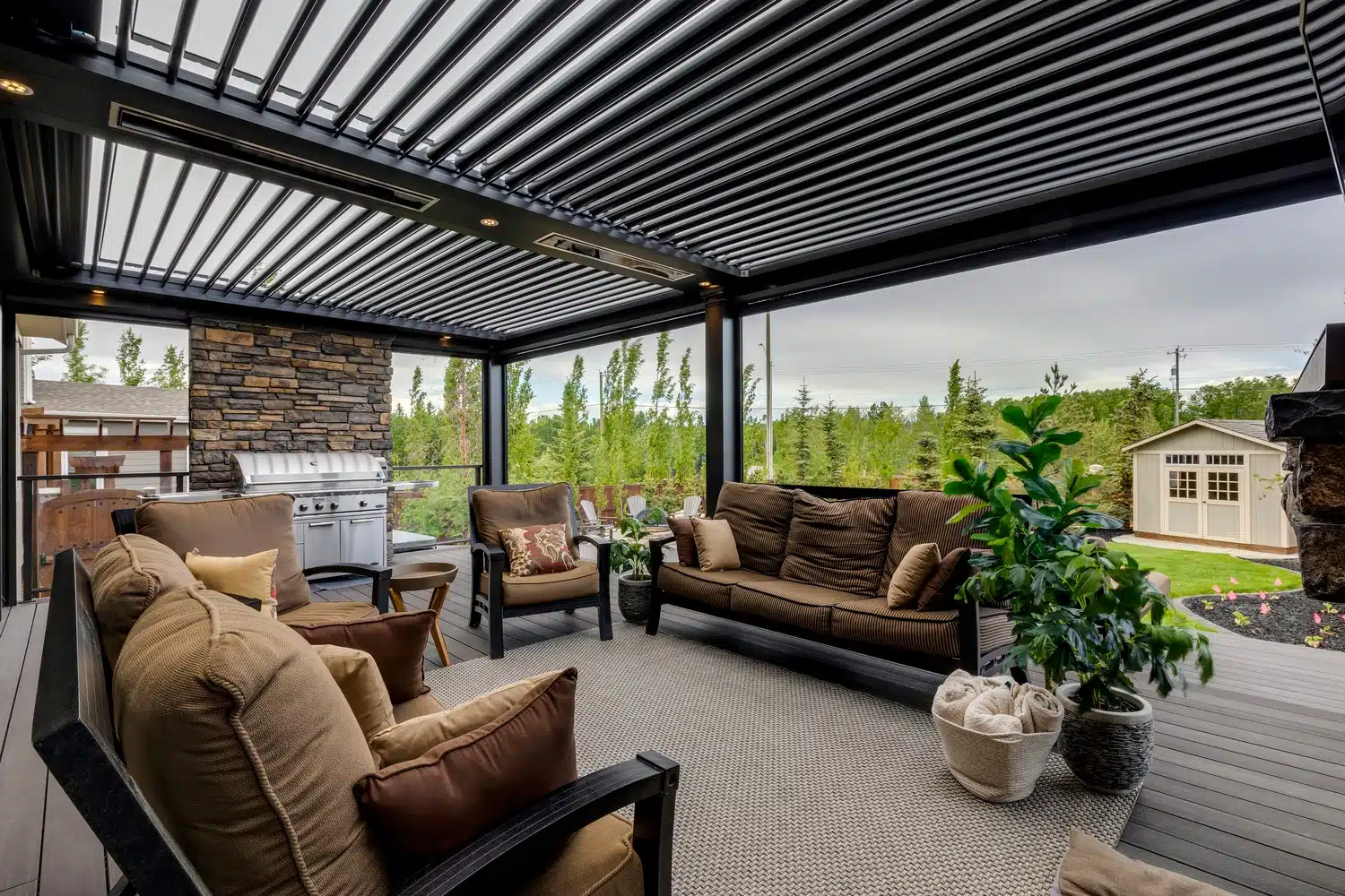 Louvered pergola covering a patio with brown cushioned seating, a stone grill area, potted plants, and a view of a green backyard with a small shed.