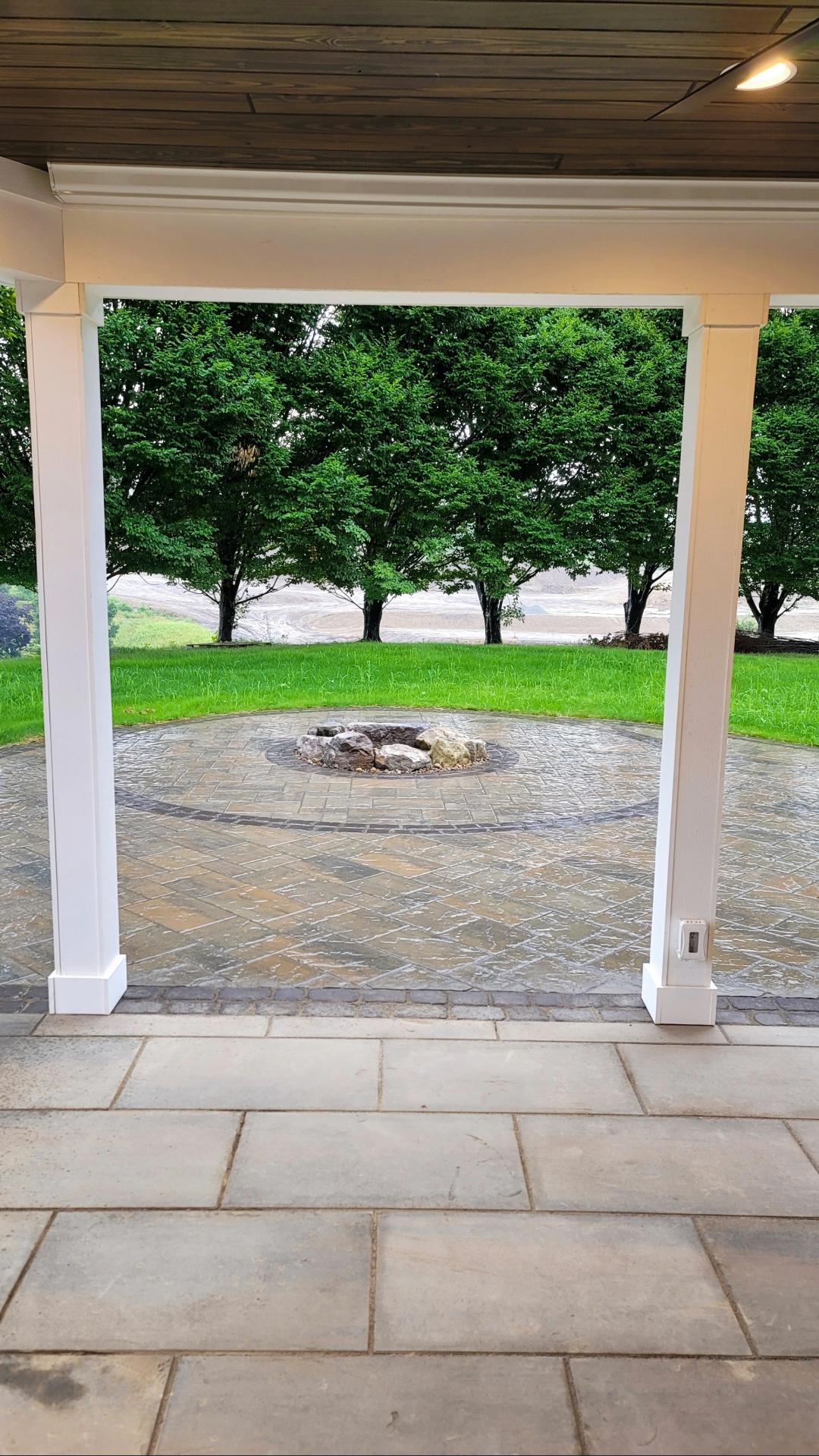 Covered patio with stone tile floor overlooking a circular stone fire pit surrounded by green grass and trees.