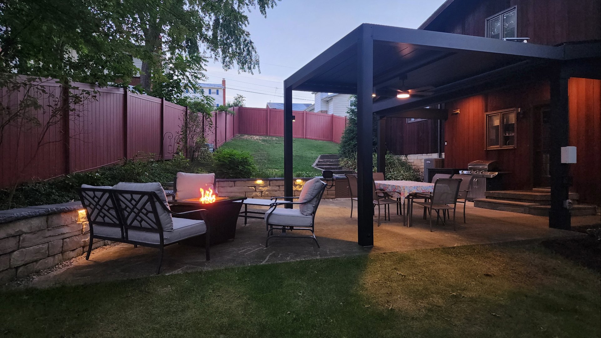 Cozy backyard patio at dusk featuring a louvered pergola around a fire pit, a covered dining area with table and chairs, and a grill near a dark wood house.