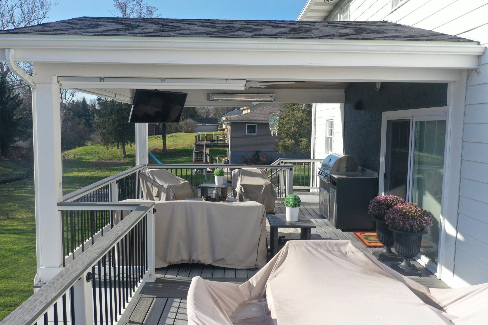 Covered patio furniture under a porch roof with a mounted TV, outdoor grill, potted plants, and a green lawn in the background.
