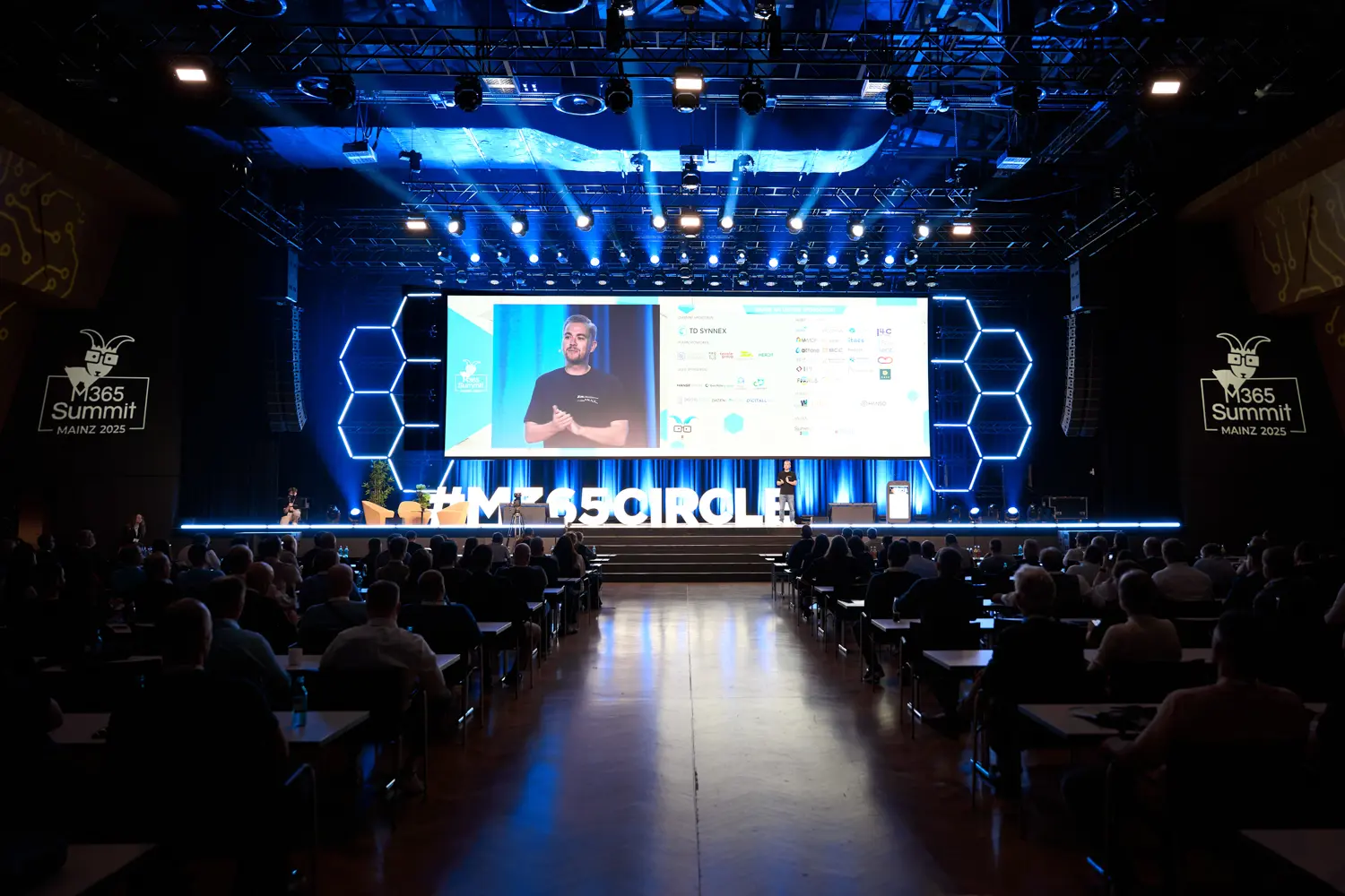 Vorlesungssaal mit Publikum bei M365 Summit Mainz 2025, Redner auf Bühne vor großer Leinwand und beleuchteten Hexagonformen.