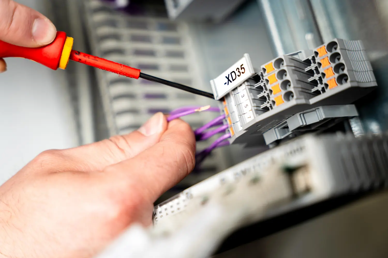 Hand hält lila Drähte und Schraubendreher mit rotem Griff zum Anschließen an eine elektrische Klemmleiste mit Beschriftung -XD35.