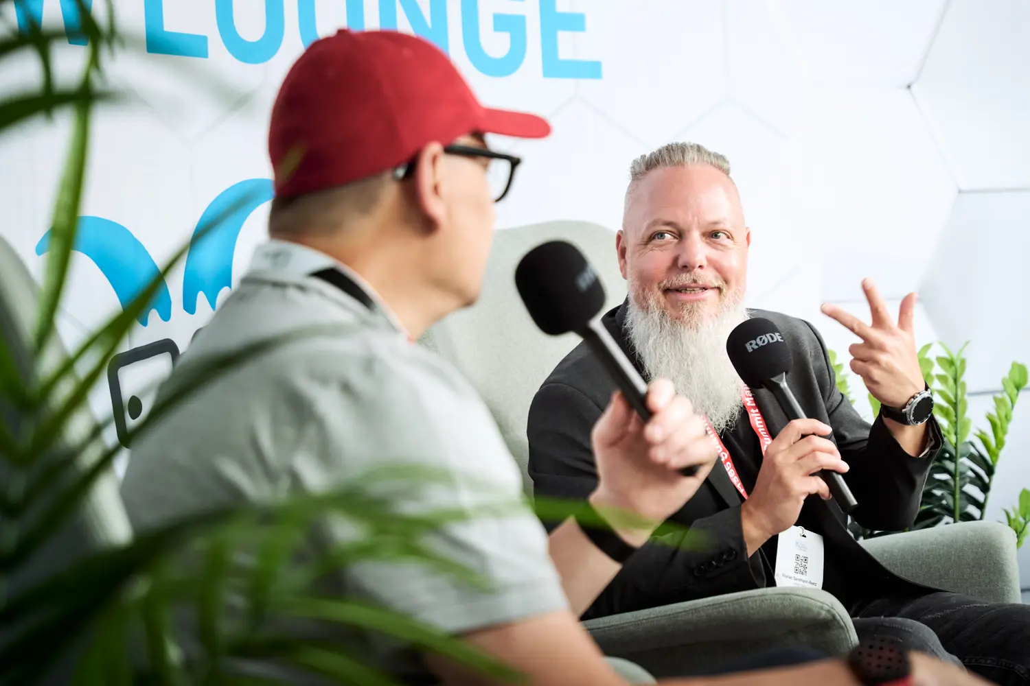 Zwei Männer führen ein Gespräch mit Mikrofonen in einem Lounge-Bereich, einer mit grauem Bart zeigt eine Geste mit den Fingern.