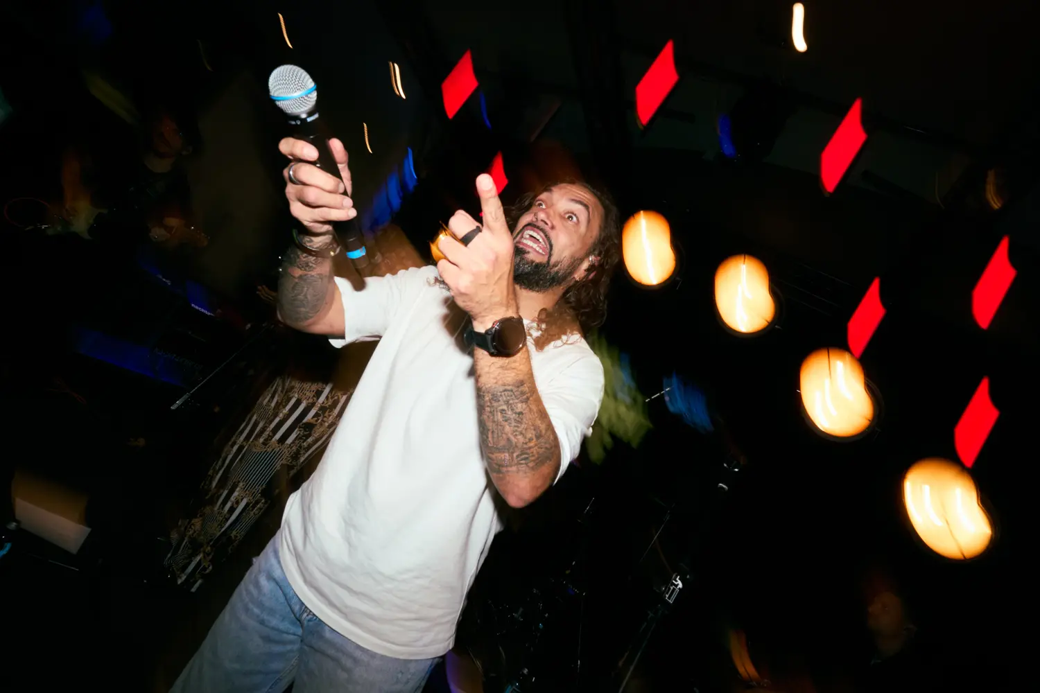 Mann mit Bart und Tattoos singt leidenschaftlich in ein Mikrofon auf einer Bühne mit bunten Lichtern.
