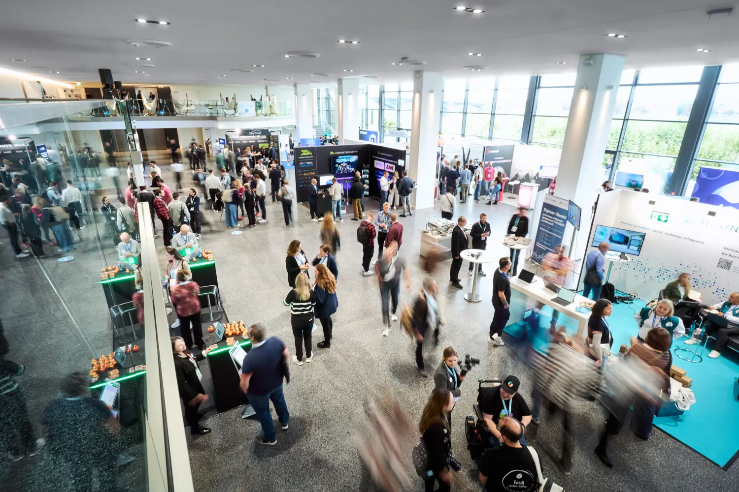 Vogelperspektive einer belebten Messehalle mit Menschen, die sich an Ständen und Tischen unterhalten und bewegen.