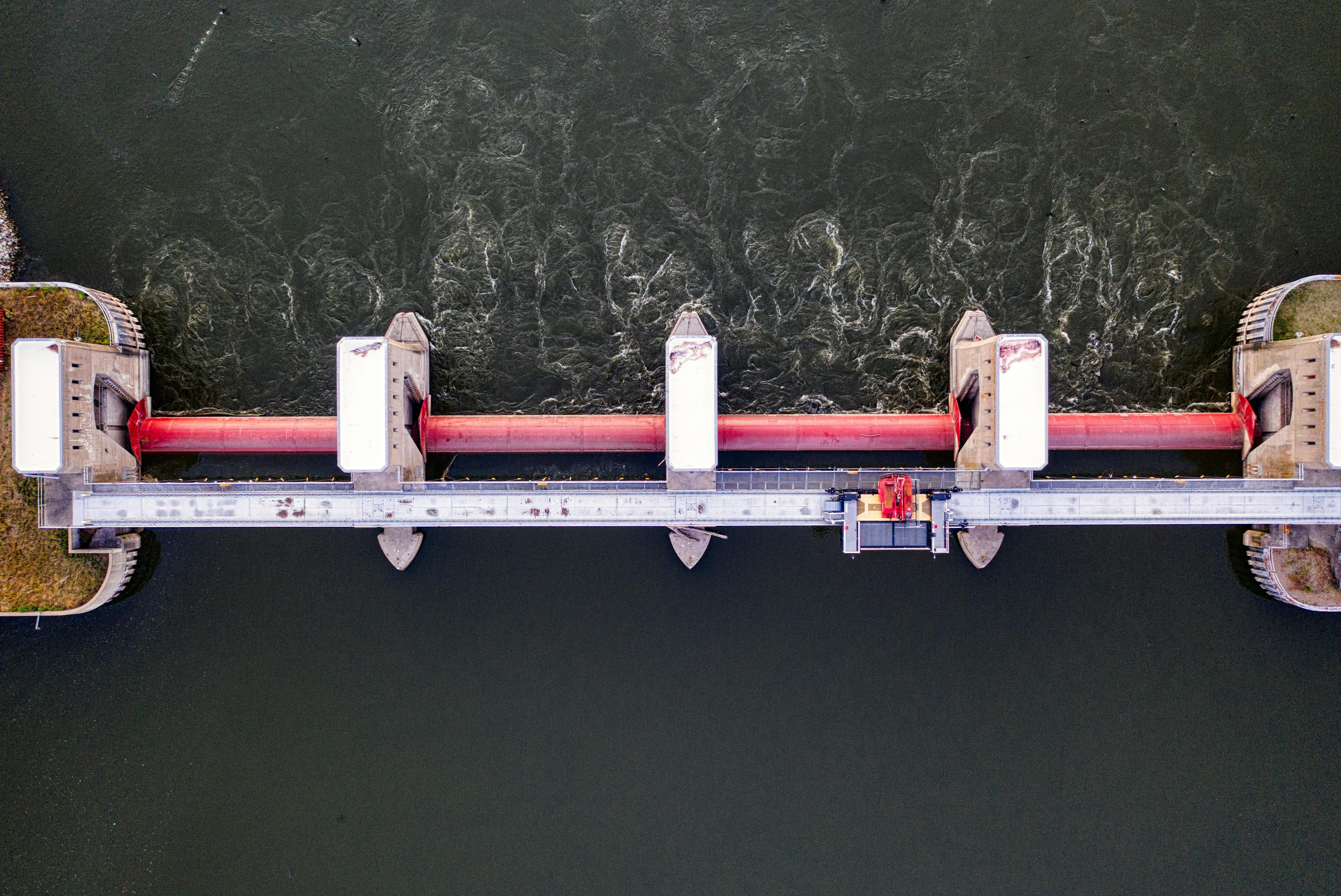 Aerial view of a dam with red gates controlling water flow from a calm reservoir to turbulent downstream.