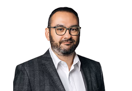 Man with dark hair, beard, glasses wearing a checkered suit jacket and white shirt.