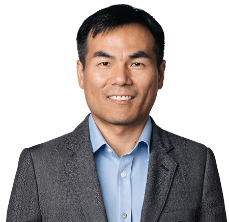 Smiling man with short black hair wearing a blue shirt and gray blazer against a white background.