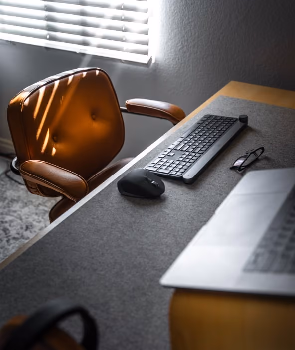 Modern workspace with a brown leather chair, computer keyboard, mouse, glasses, and a laptop on a wooden desk near a window with blinds.