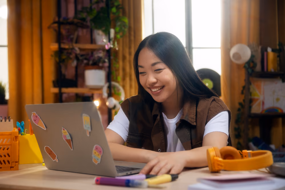 Smiling young woman with long dark hair working on a laptop decorated with ice cream stickers.