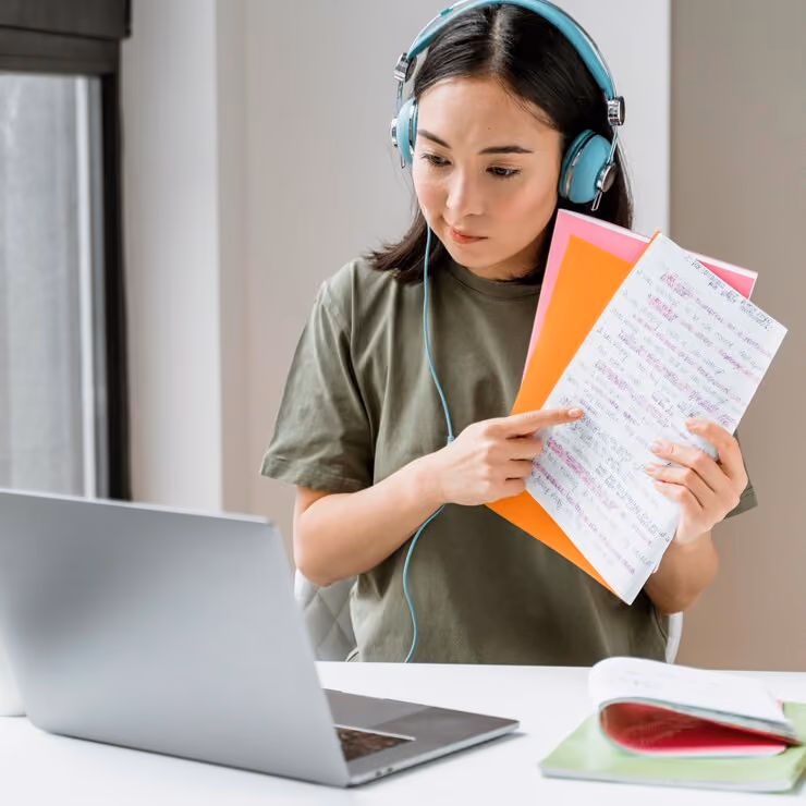 Young woman wearing headphones pointing to notes while video calling on a laptop.