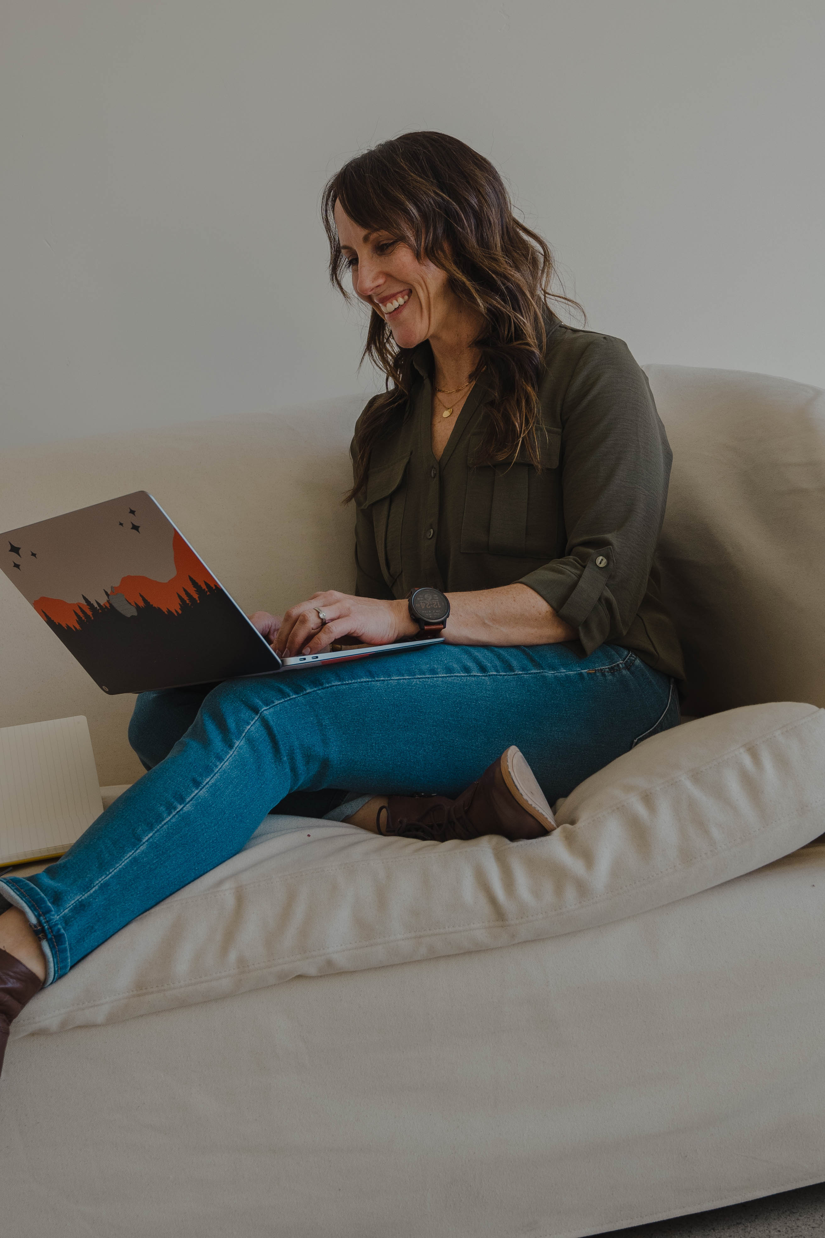 Andrea Giachetti FNP, CFMP in a green shirt and jeans sitting cross-legged on a beige couch using a laptop with a mountain and forest decal.