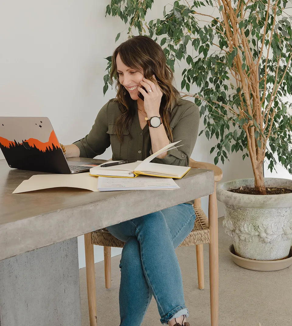 Andrea Giachetti FNP, CFMP sitting at a table with a laptop, open notebook, and papers, talking on a phone with a potted plant in the background.