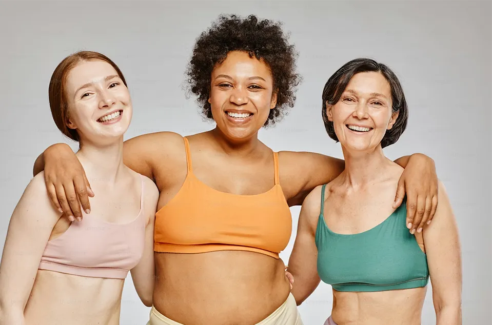 Three diverse smiling women in sports bras standing close with arms around each other against a plain background.