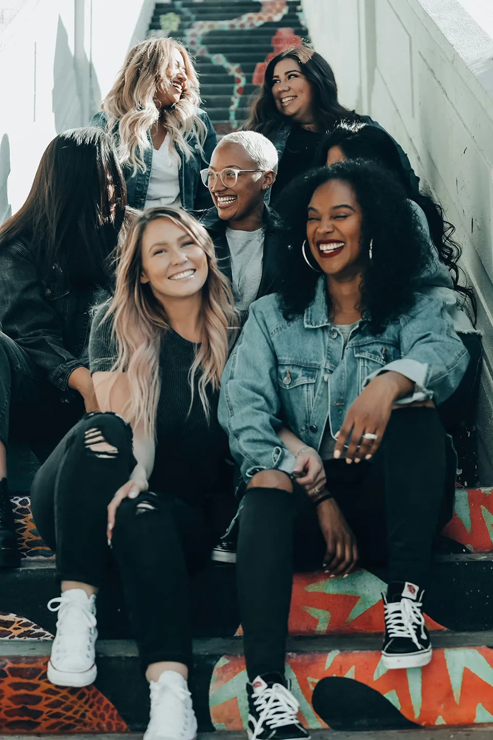 Group of six diverse women sitting on colorful painted stairs, smiling and laughing together.