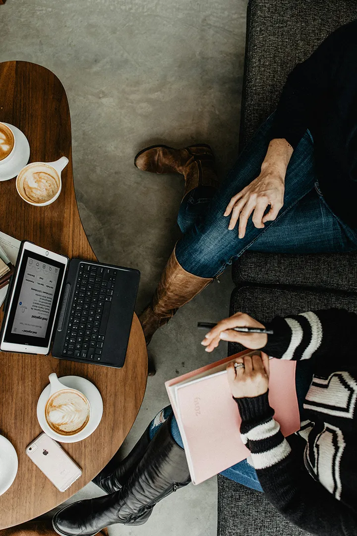 Two people sitting on a couch with a wooden coffee table holding lattes, a tablet with keyboard, a phone, and a planner.