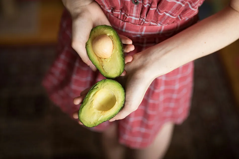 Person in a red checkered dress holding two halves of a ripe avocado, one with the pit and one without.