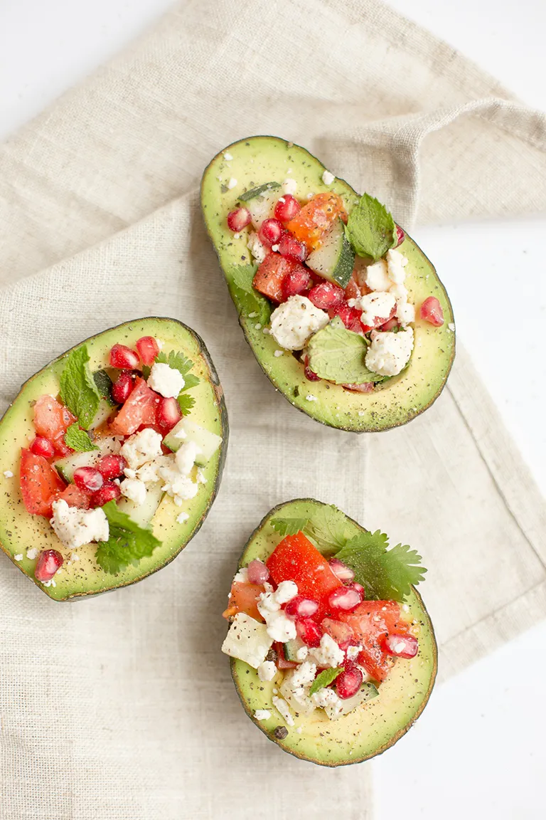 Three avocado halves filled with diced tomato, cucumber, feta cheese, pomegranate seeds, and fresh herbs on a beige cloth.