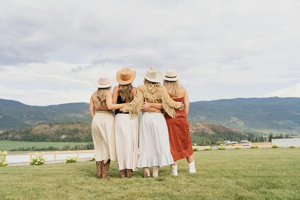 Four women standing on grass with their backs to the camera, wearing skirts, hats, and boots, overlooking a mountainous landscape.