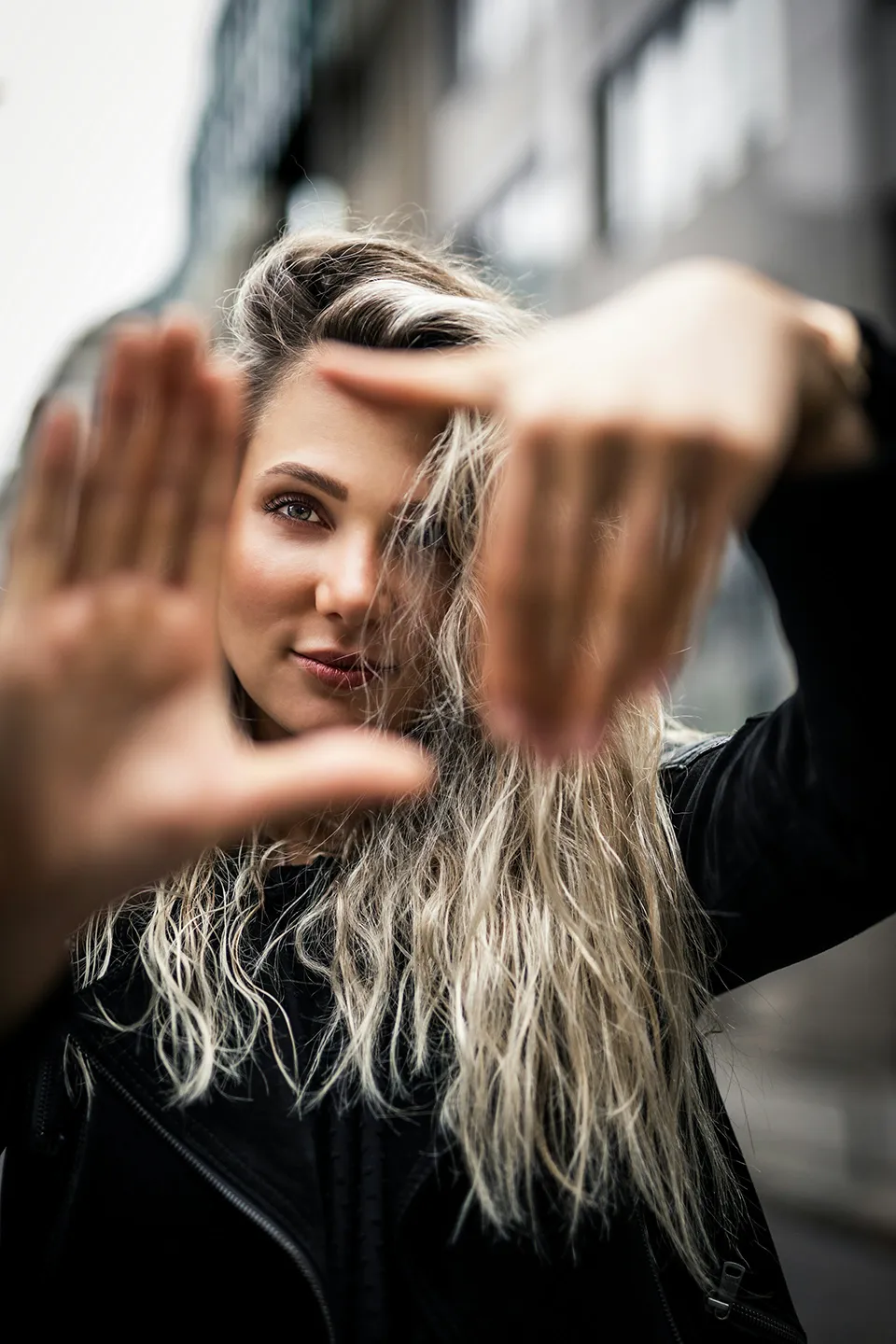 Young woman with long blonde hair framing her face with her hands, creating a rectangular shape in an urban setting.