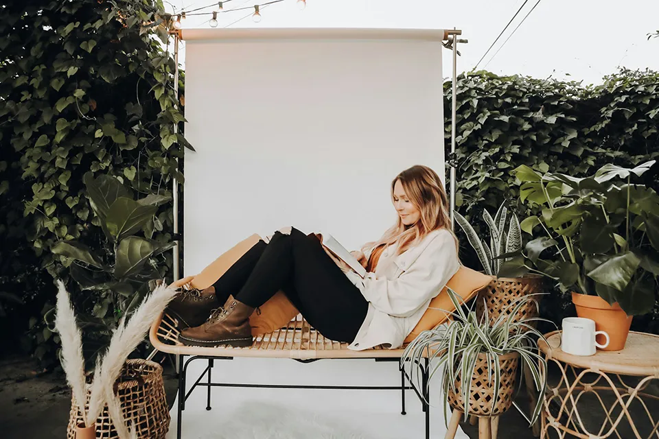Woman with light brown hair wearing a white jacket and black ripped jeans reading a book while reclining on a wicker bench surrounded by green plants.