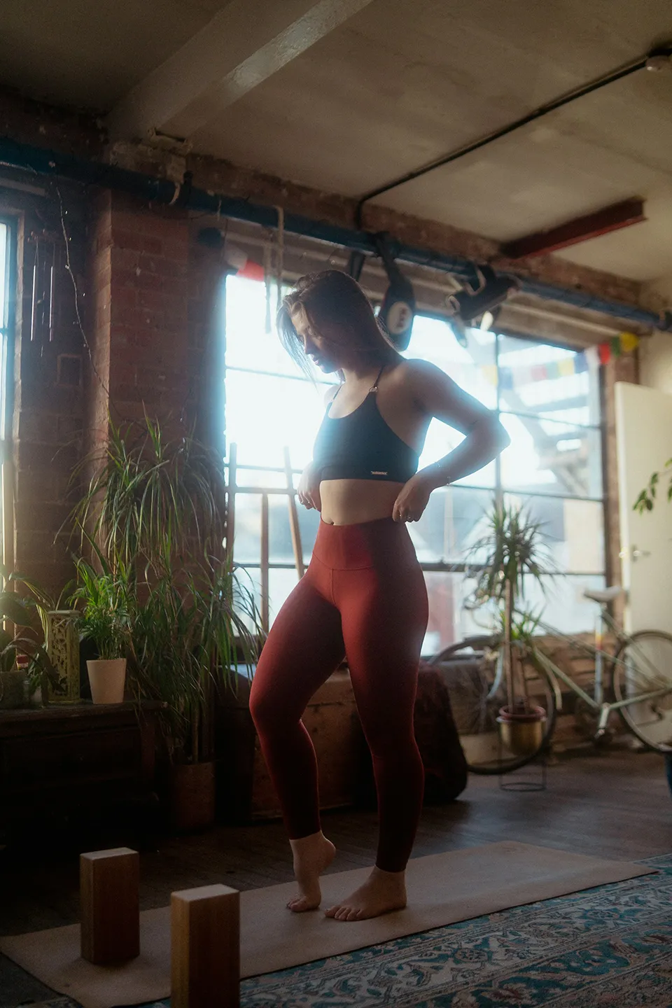 Woman in black sports bra and red leggings standing on a yoga mat in a sunlit room with plants and a bicycle.