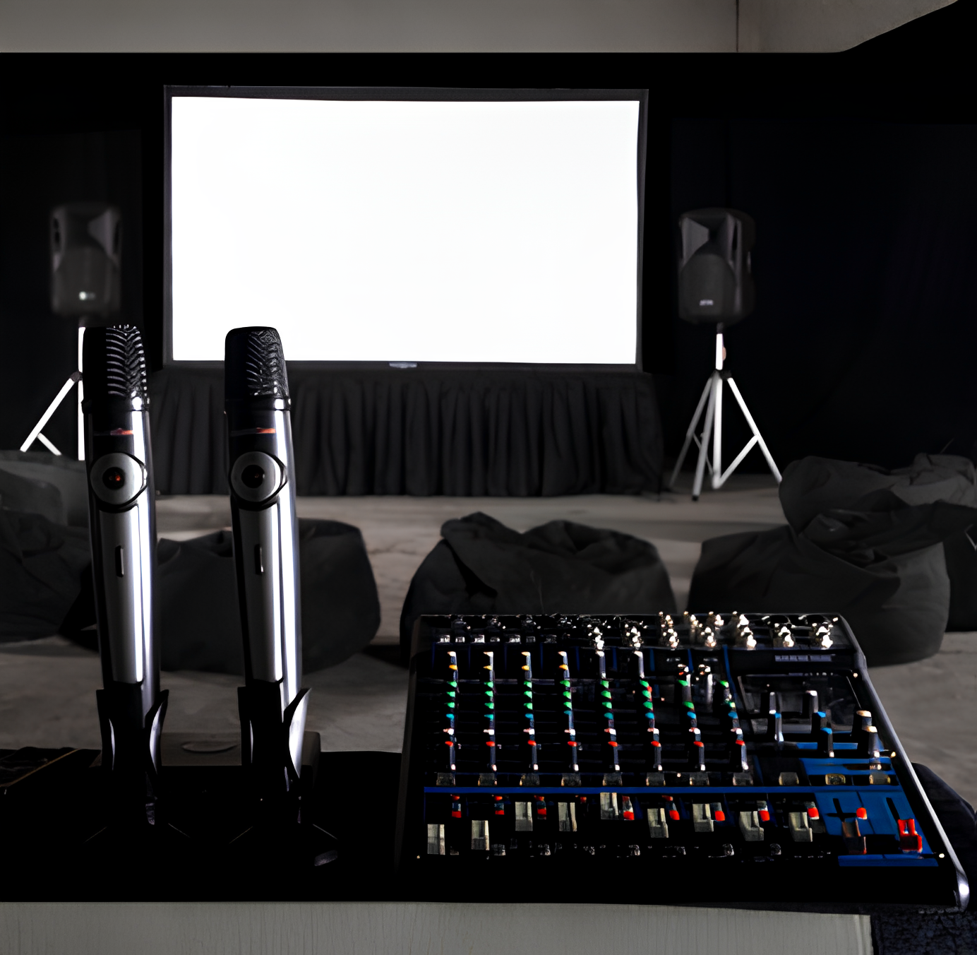 Two wireless microphones and an audio mixer on a table facing a blank projection screen with black bean bags and speakers in the background.