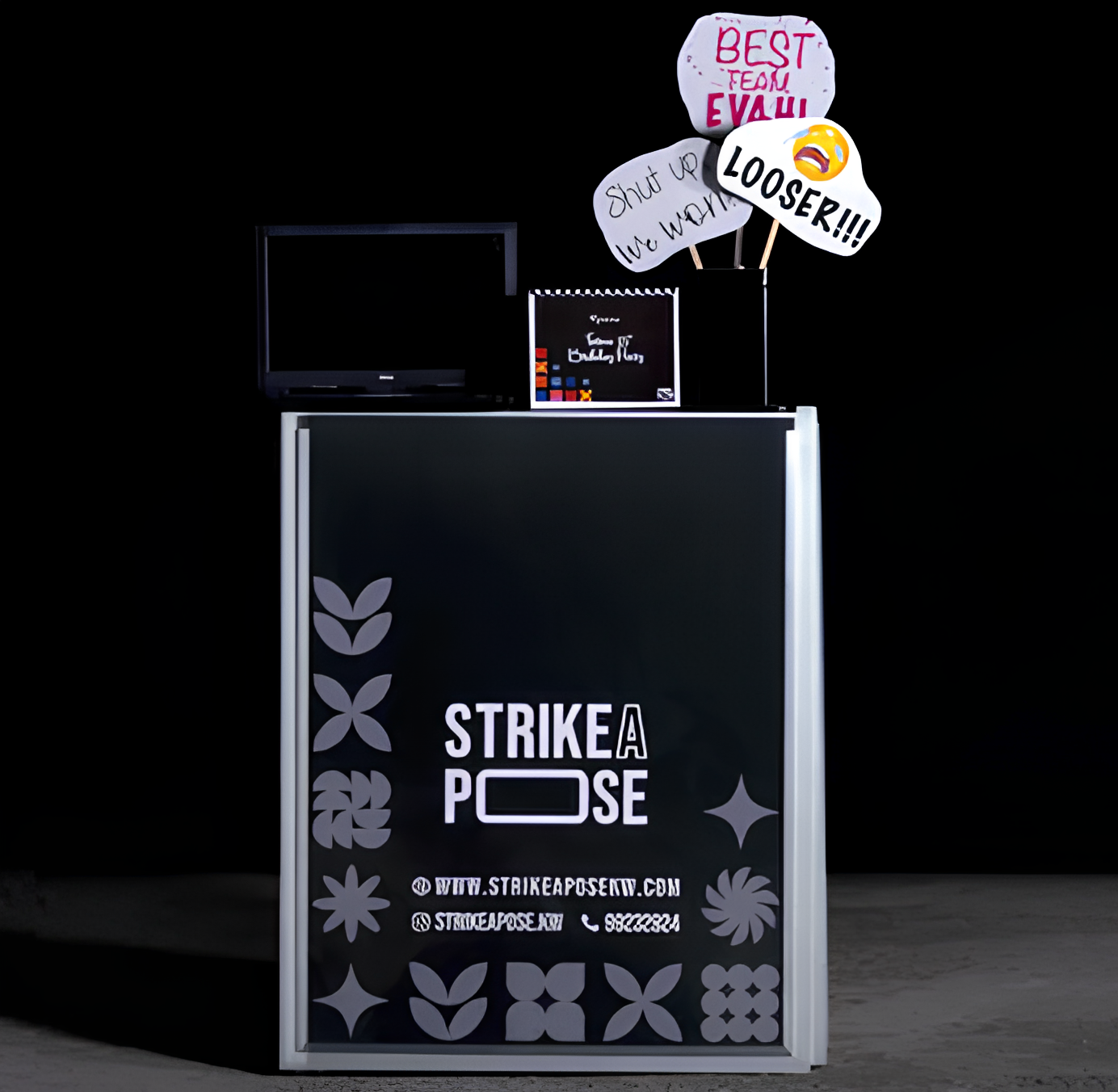 Photo booth setup with a black sign reading 'Strike A Pose' surrounded by floral patterns and a small monitor on top, alongside props with phrases like 'Best Team Evah', 'Shut up we work', and 'Looser!!!' emoticon.
