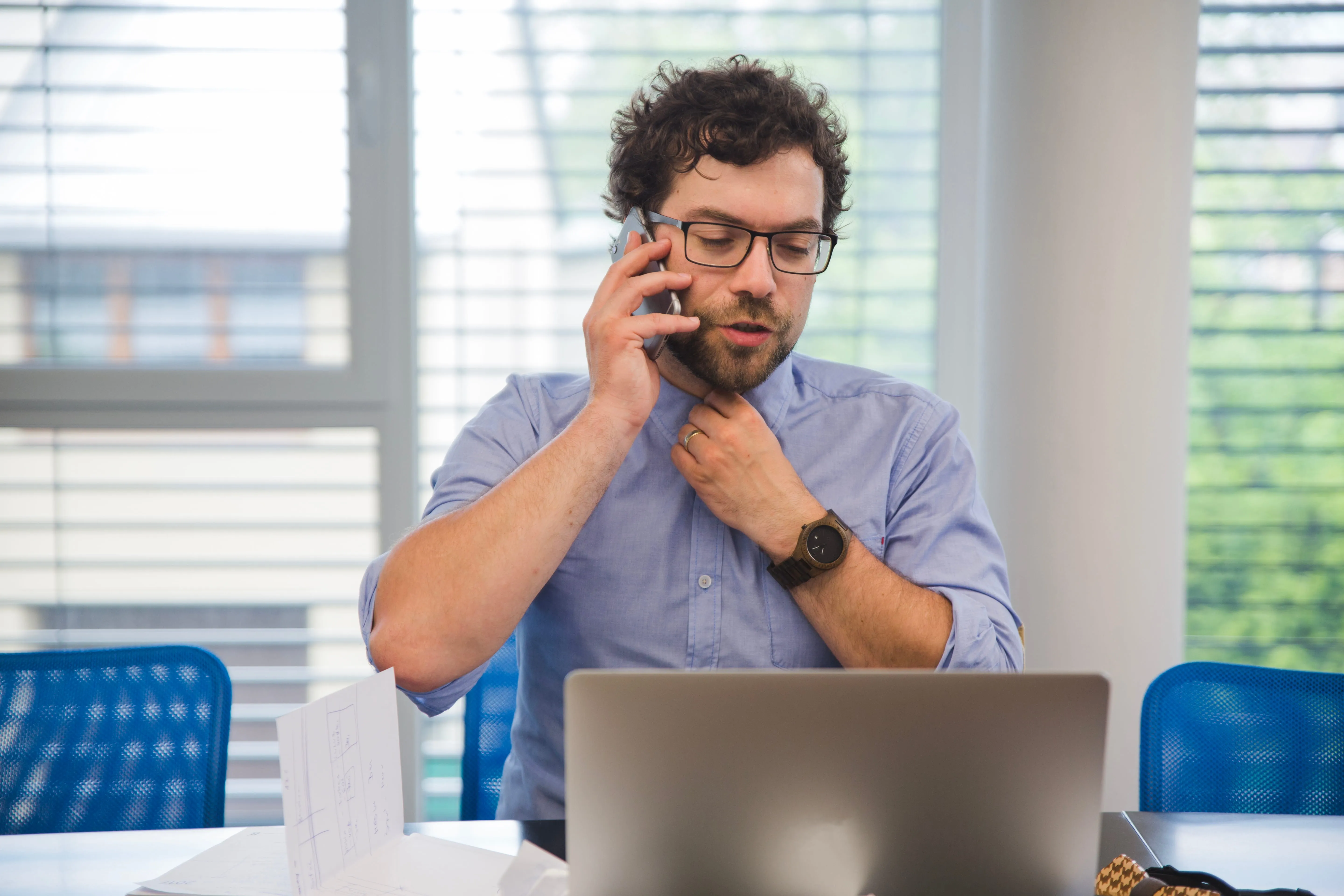 Man in a blue shirt talking on a smartphone while working on a laptop in a modern office.
