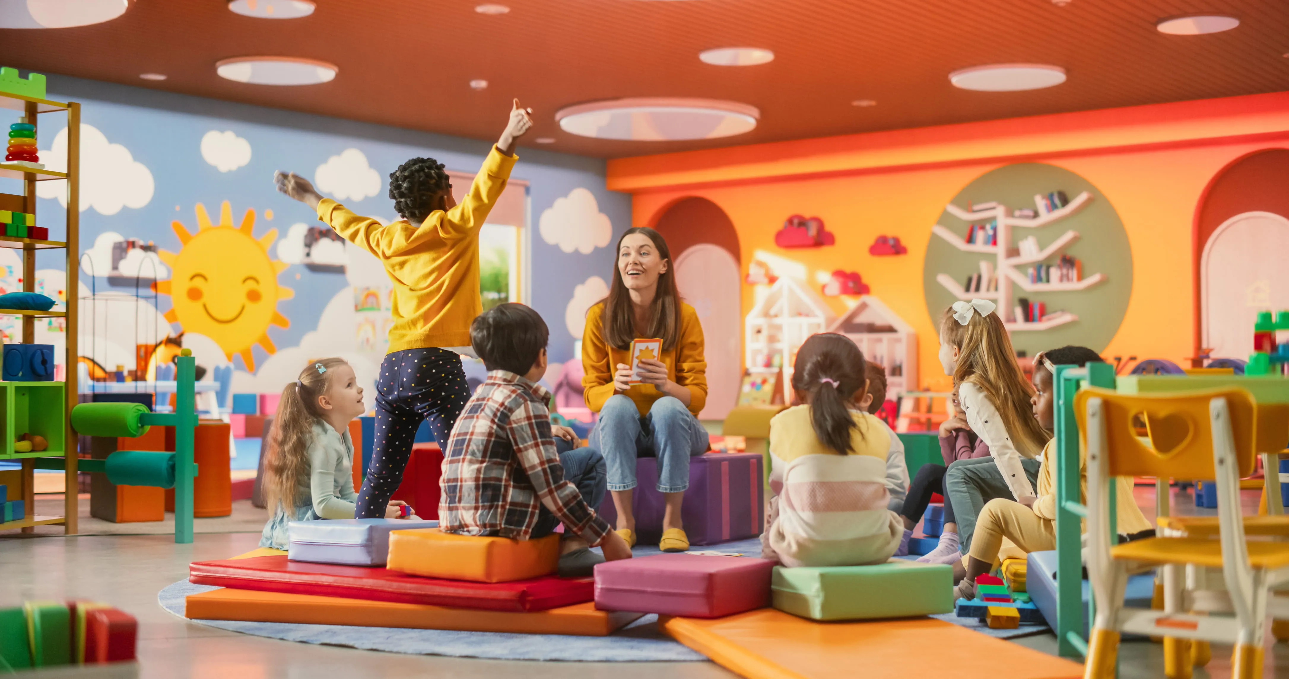 Teacher with children sitting on colourful cushions in a lively classroom decorated with sun and cloud murals.