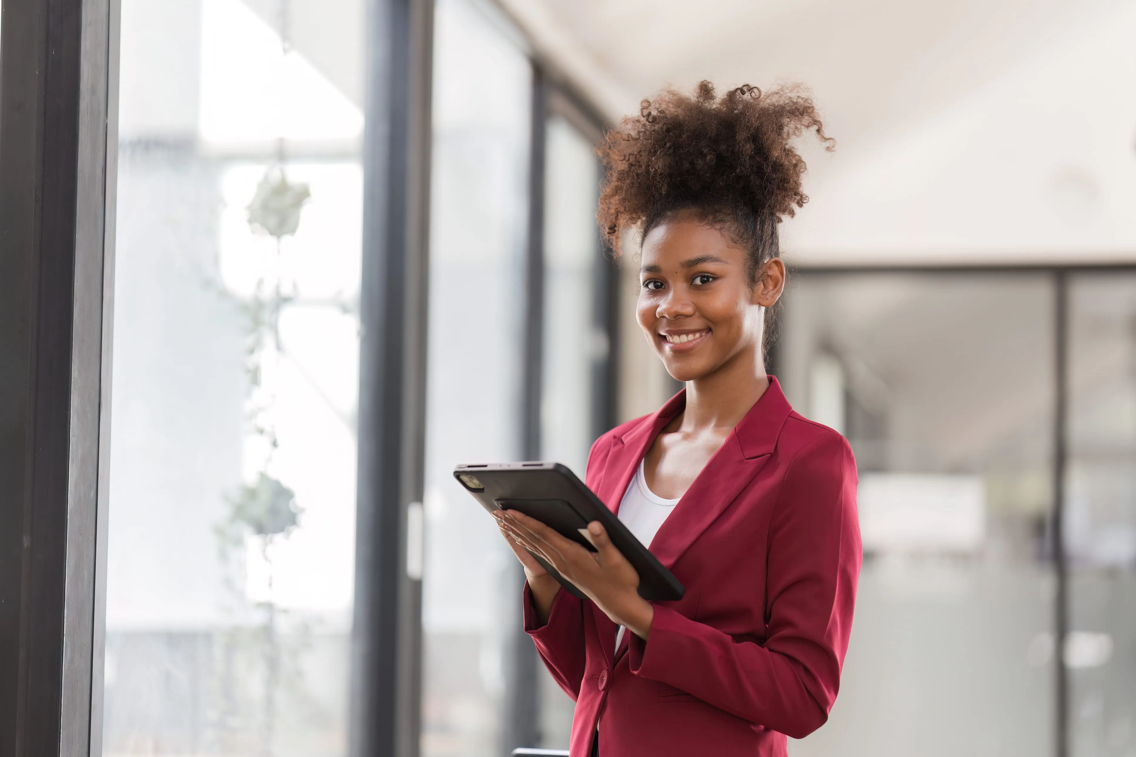 Young woman in a red blazer smiling and holding a tablet in a modern office.
