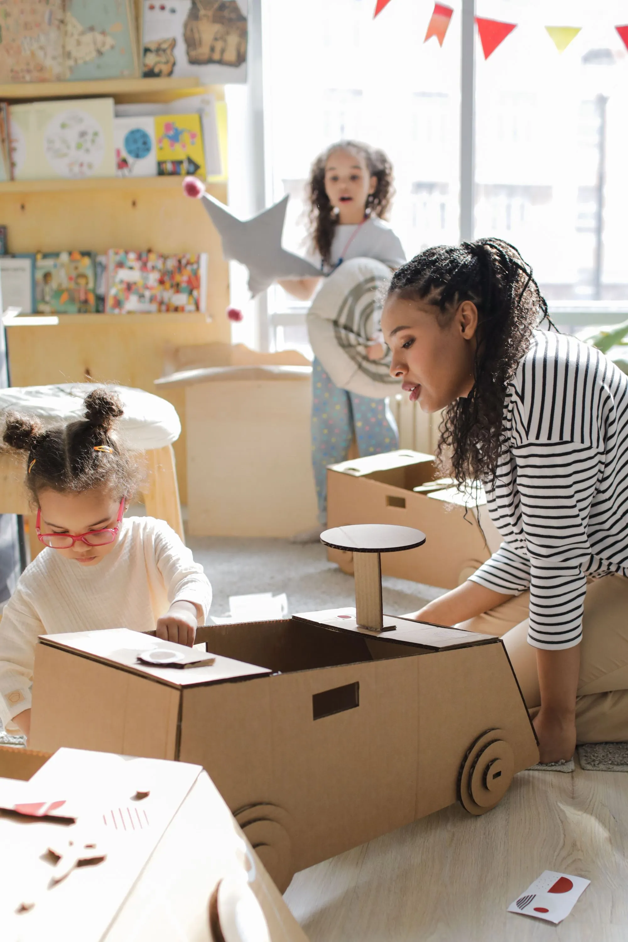 Young girl in glasses building a cardboard car with an educator in a bright childcare classroom