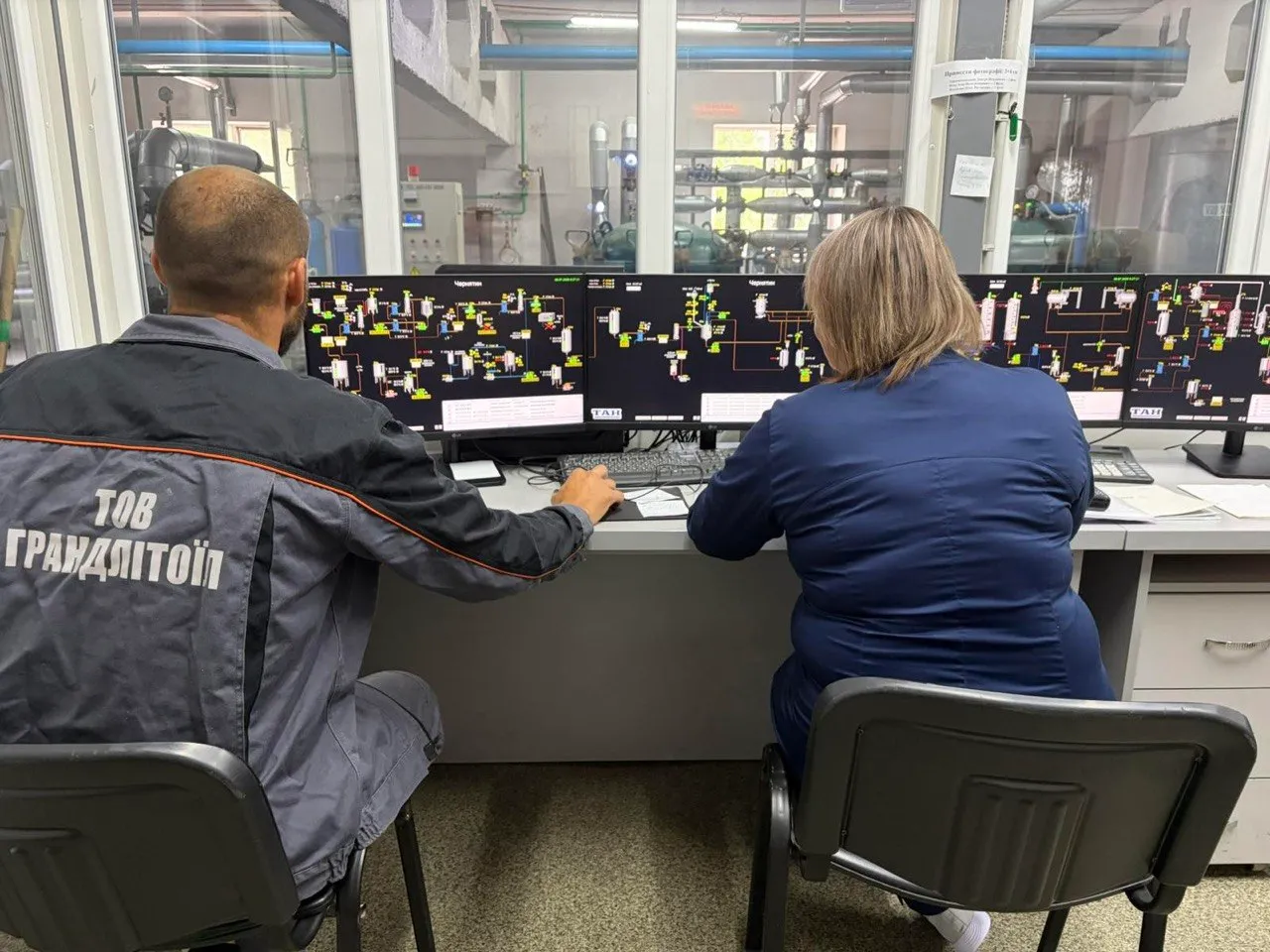Двоє працівників у робочій формі сидять перед трьома екранами з технічною схемою.