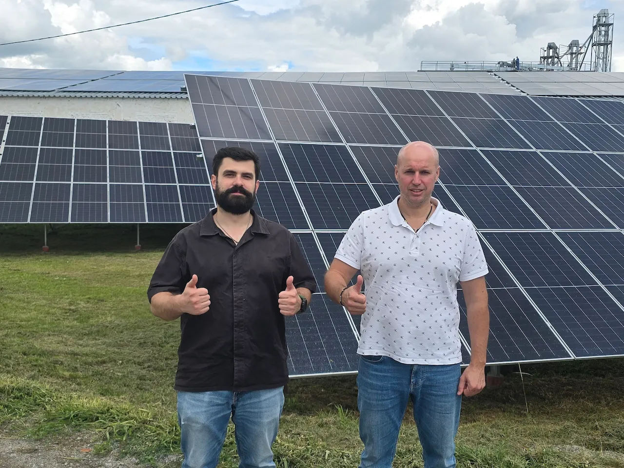 Два чоловіки стоять із піднятими великими пальцями на тлі сонячних панелей на зеленій траві.