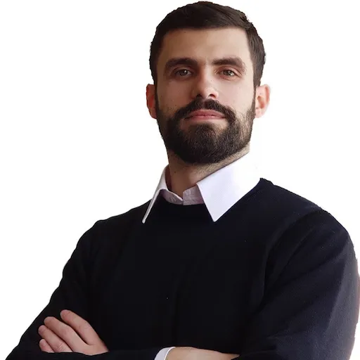 Чоловік із бородою в чорному светрі та білій сорочці, зі складеними руками, дивиться вперед.