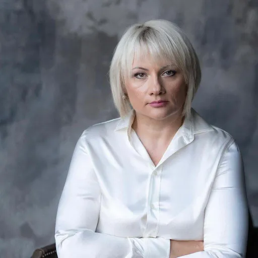 Жінка з коротким світлим волоссям у білій сорочці на сіруватому фоні з серйозним виразом обличчя.